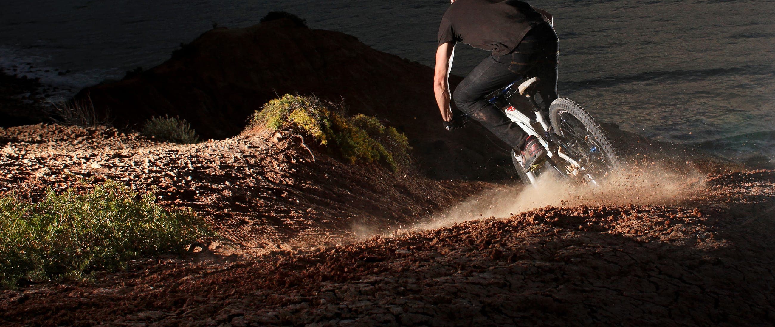 A mountain biker railing a berm of red dirt above the ocean in low afternoon light.