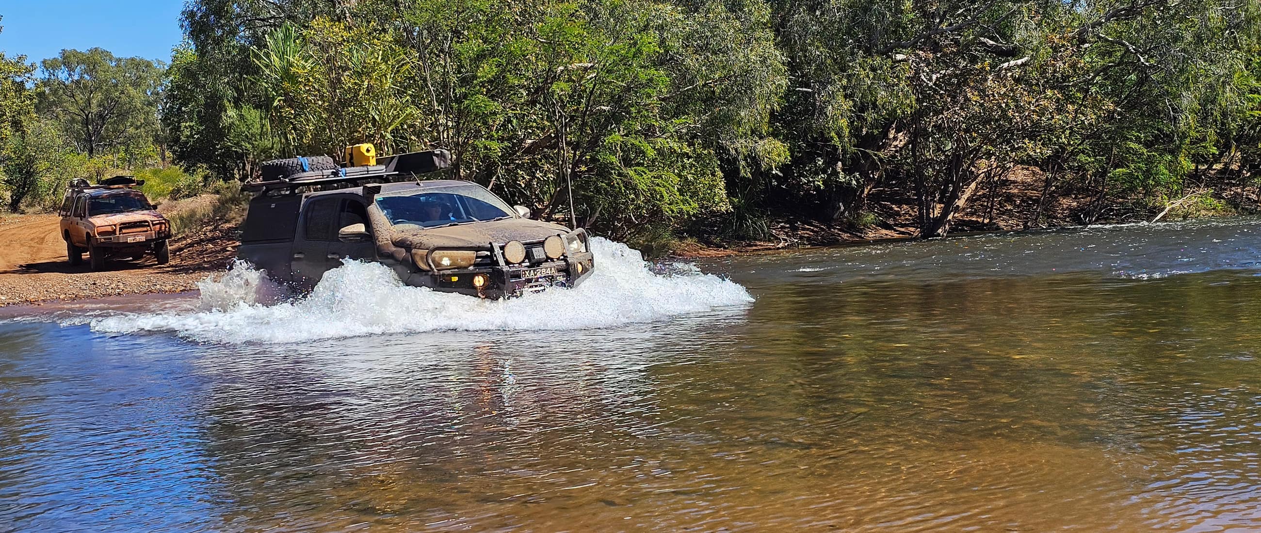 A loaded four wheel drive pushing a bow wave through a river crossing while a second vehicle waits on the bank.