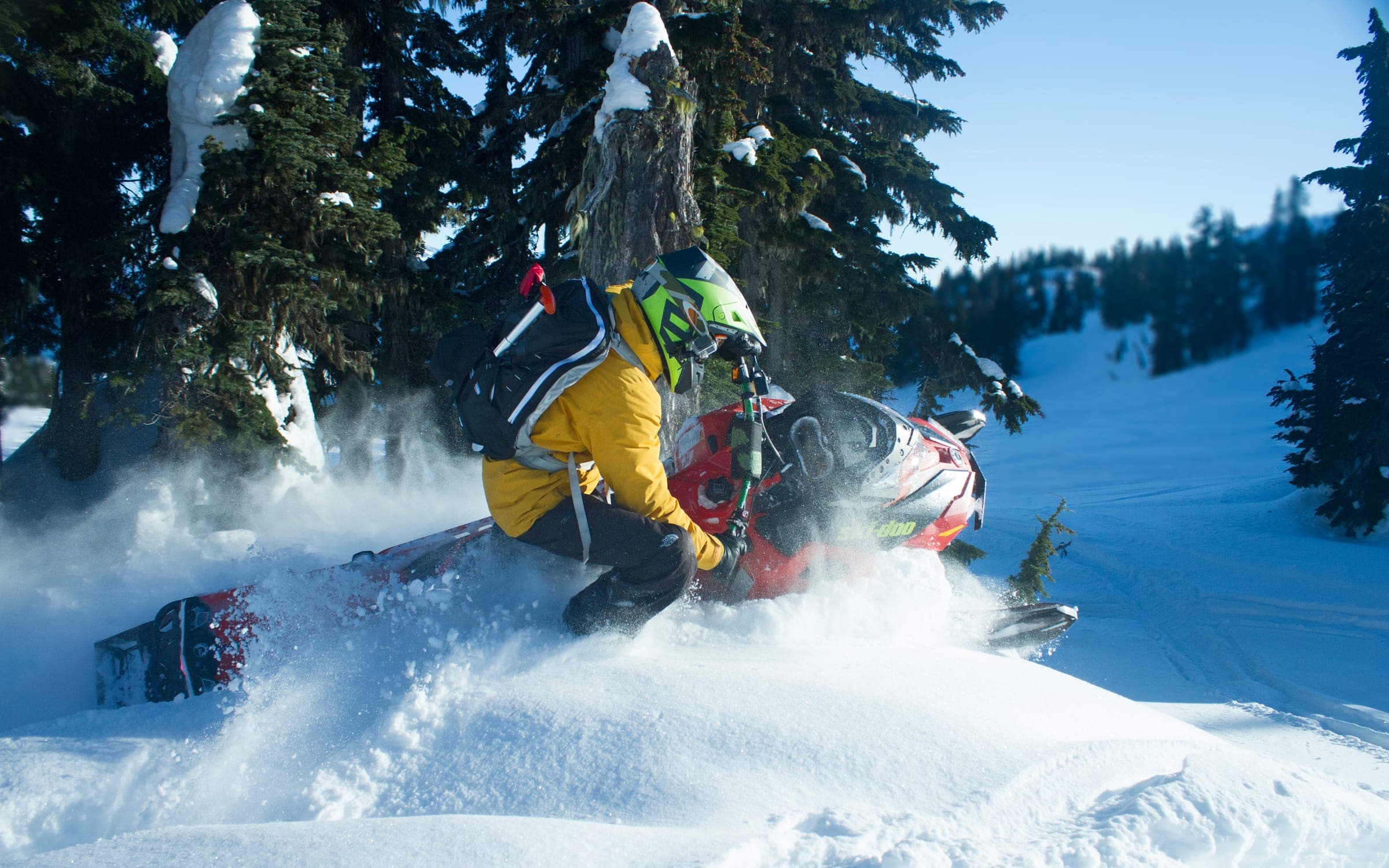 A snowmobile laid hard over in deep snow, track spraying, sunlit timber behind.